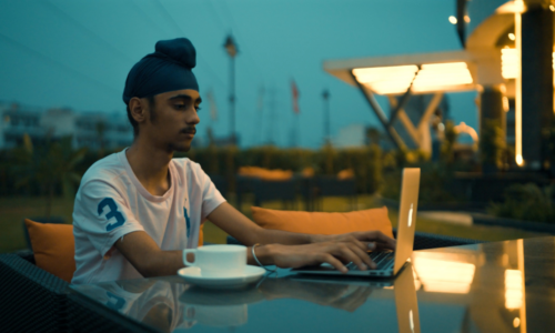 A young person sits at a table outside and uses a laptop. 