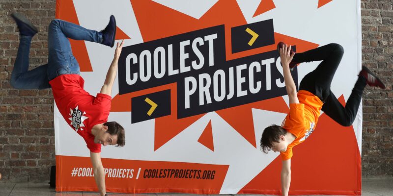 A Coolest Projects sign with two people doing handstands in front of it.
