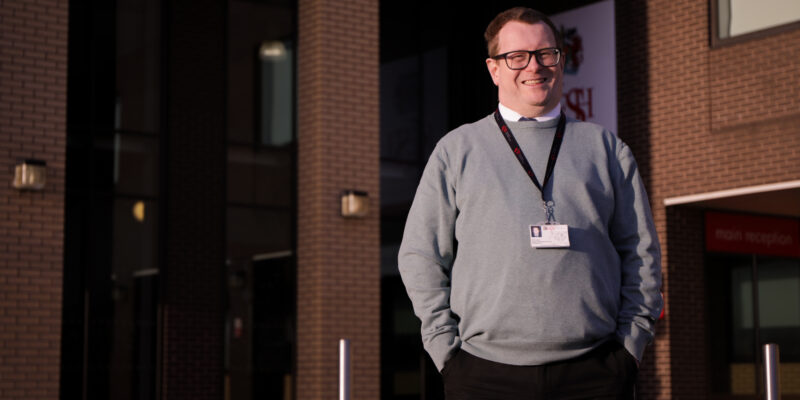 A smiling computer science teacher stands in front of a school building.